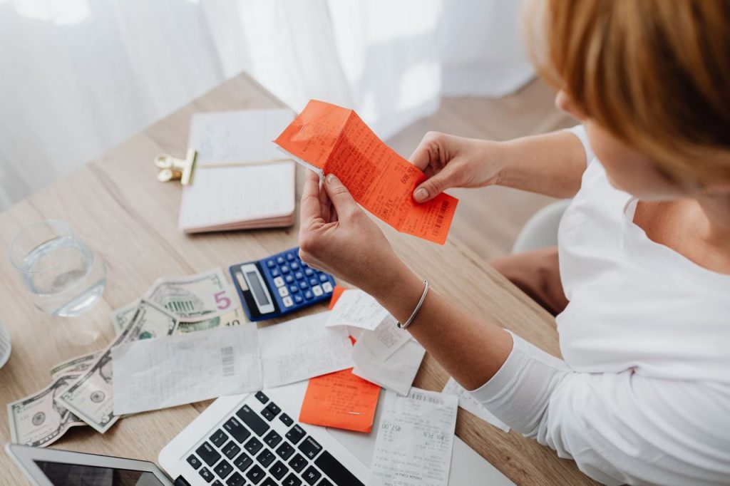 Conta de luz e passagens aéreas ajudam a moderar a inflação em outubro Mulher usa uma calculadora e analisa recibos em uma mesa de trabalho, em uma cena de controle financeiro doméstico.