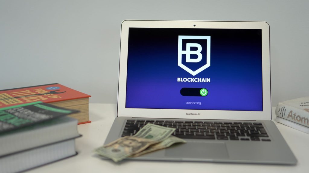 Notebook exibe uma tela sobre conexões de blockchain sobre uma mesa com livros e cédulas de dólar.